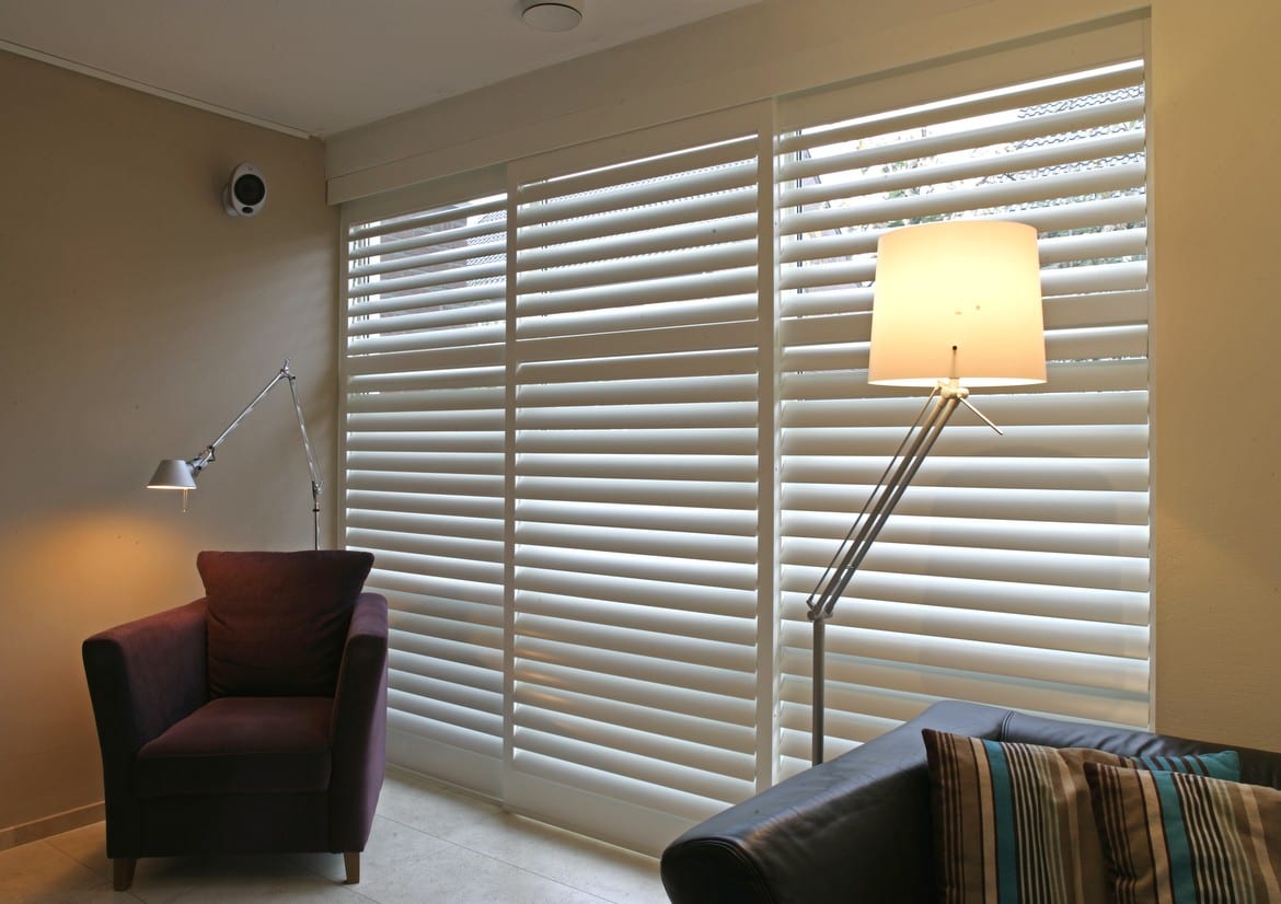By-pass tracked shutters in a living room with a lamp and armchair