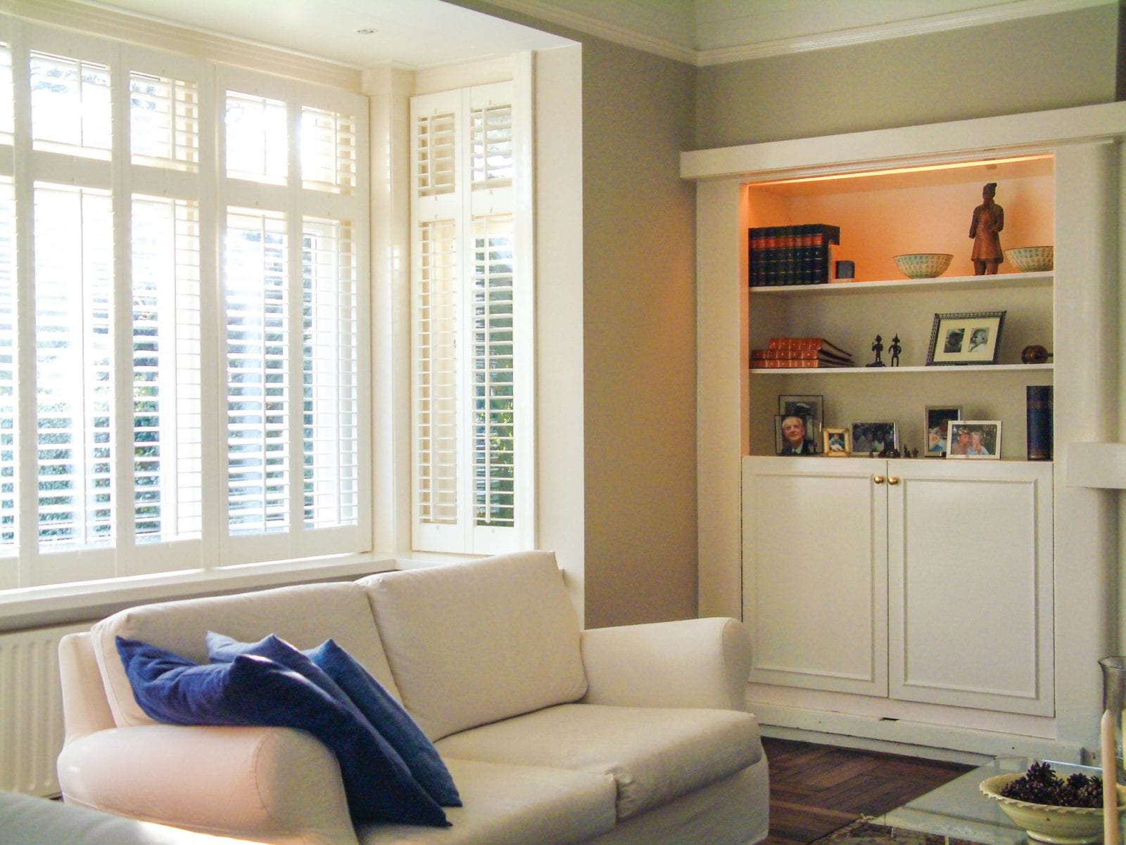 White shutters on a square bay window in a living room with a cream sofa and built-in shelving