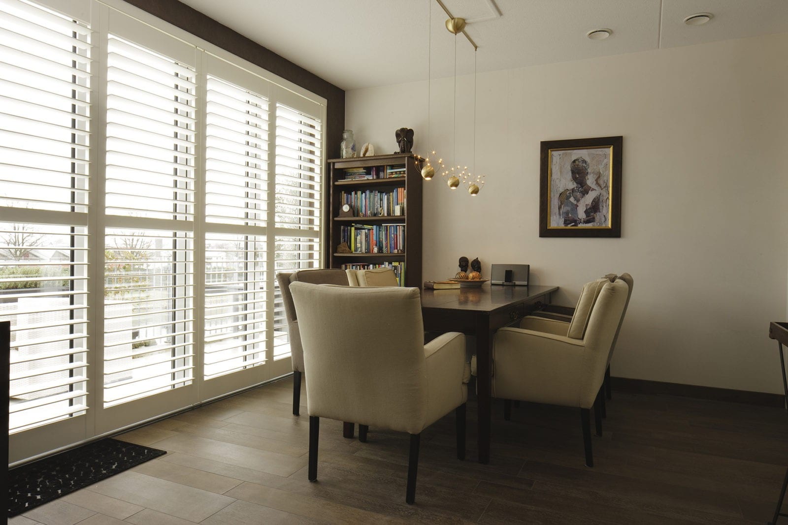 Tracked shutters across windows in a dining room with a table and bookcase