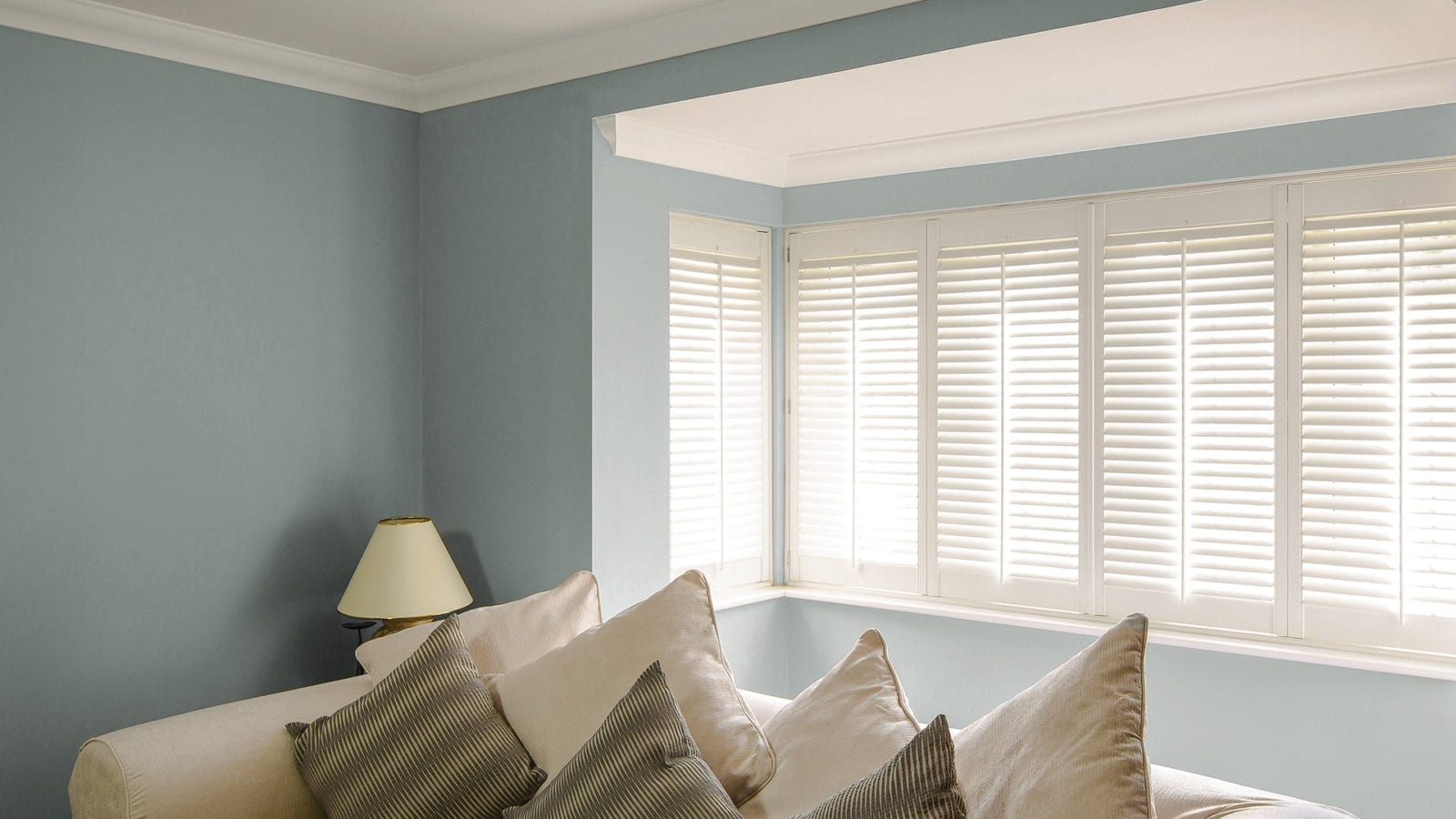 White bay window shutters in a blue-grey living room with striped cushions