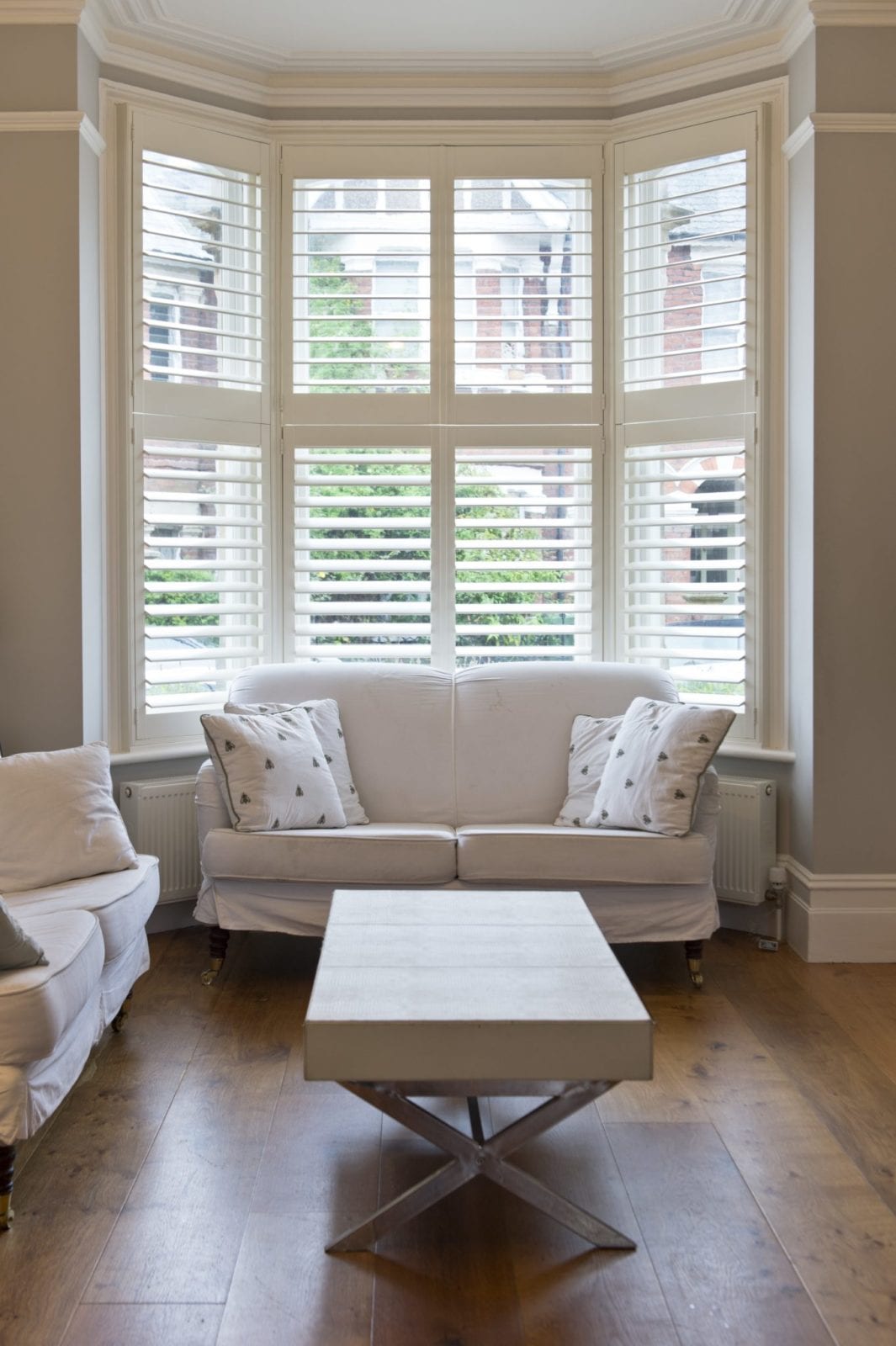 Tall white bay window shutters in a lounge with bookshelves and patterned curtains