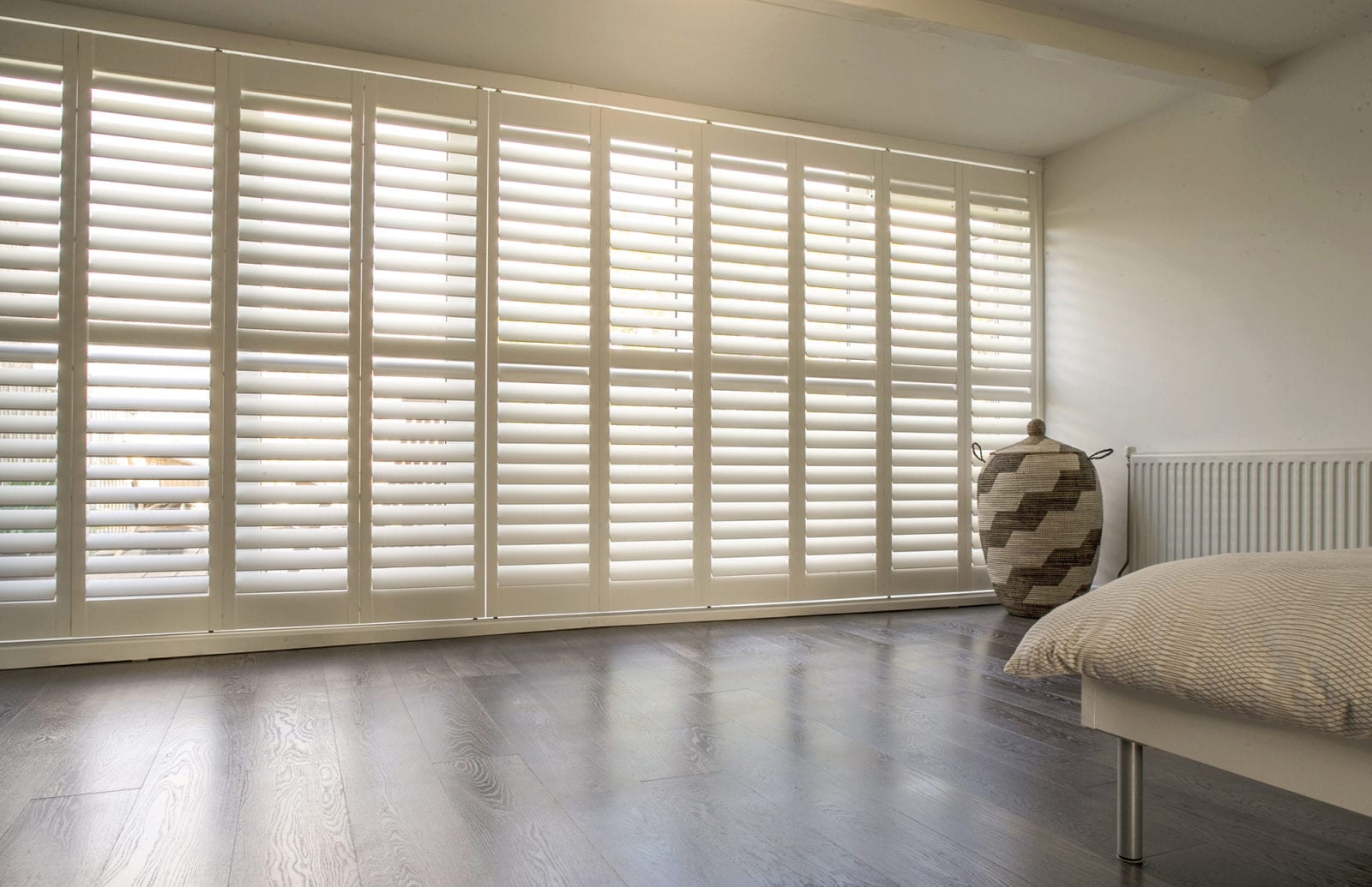 Wide white tracked shutters across a bedroom window wall above a wooden floor