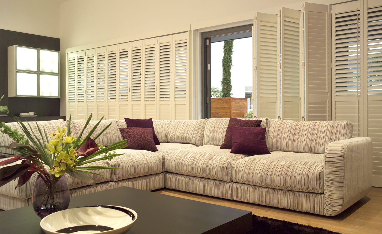 Tracked shutters folded open beside a patio door in a living room with a striped sofa